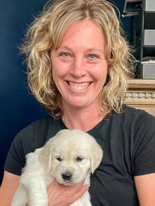 Before color analysis, a woman with blonde hair wearing a black top while holding a puppy. Her skin appears flat and the black overwhelms her natural coloring.